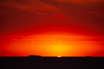 Cargo ship on the sea in sunset with cloud formations in red light. Sun touches the sea and is almost gone.