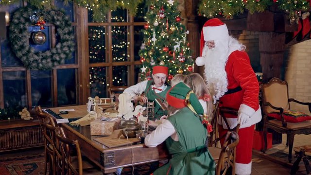 Santa Claus, elves in green costumes and two kids preparing gifts and letters for holiday. Elves and children are sitting at the wooden table on the background of christmas decorations.