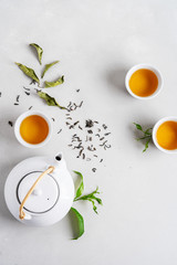 Tea concept with three white cups of tea and teapot surrounded with fresh green tea leaves and dry leaves,  top view composition on concrete background with copy space.