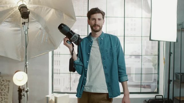 In the Photo Studio with Professional Equipment: Portrait of the Handsome Photographer Holding State of the Art Camera Ready to Take Pictures with Softboxes Lighting in Background