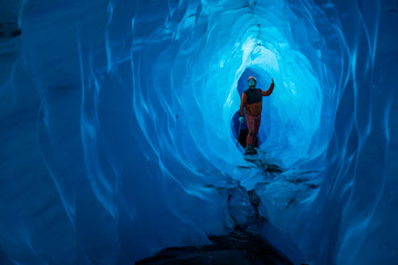 Ice climber in all orange looking up at narrow section of an ice cave. Light shines through the...