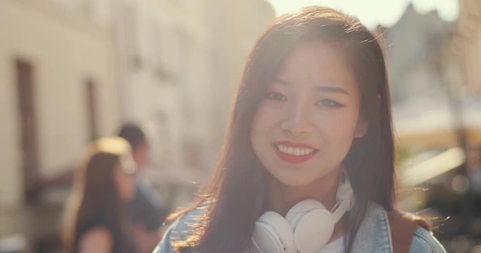Portrait of the beautiful Asian young woman with big whit eheadphones on her neck turning her dace and smiling cheerfully to the camera in sunlight while walking as a tourist. Close up.