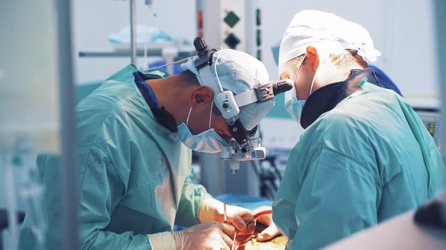 Surgeon with magnifying glass on his head is working with assistants in operation room. Team of specialists in medical uniform performing an operation to a patient with special instruments in clinic.