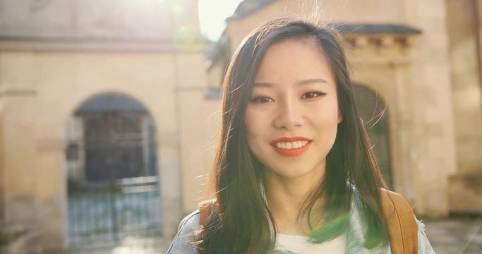 Portrait of the beautiful Asian young woman with red lips turning her face to the camera and smiling happily while having sightseeing roung the city on a sunny day. Close up.
