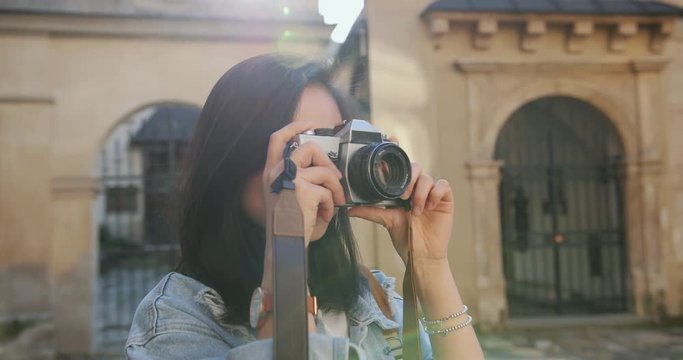 Close up of the young pretty brunette asian woman taking a photo with a photocamera while doing sightseeing in the historical part of the city where she walking on a summer day.