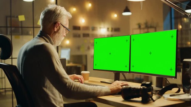 Creative Middle Aged Designer Sitting at His Desk Uses Desktop Computer with Two Green Mock-up Screens. Professional Office Employee Working Late in the Evening in His Studio 