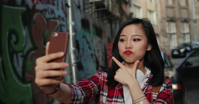 Beautiful and cute Asian young woman in headphones and motley red shirt smiling cheerfully and posing to the smartphone camera while taking selfie photos at the graffity wall outdoors.