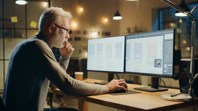 Creative Middle Aged Mobile Software Designer Sitting at His Desk Uses Desktop Computer with Two Screens Showing Smartphone Application Design Process. Stylish Modern Studio Office