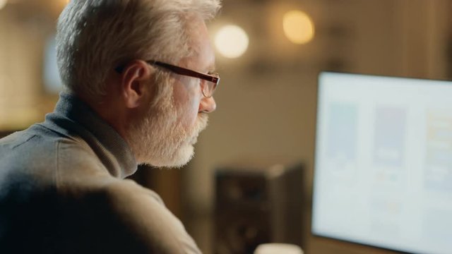 Creative Middle Aged Mobile Software Designer Sitting at His Desk Uses Desktop Computer with Two Screens Showing Smartphone Application Design Process
