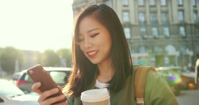Close up of the smiled Asian young beautiful woman typing a message on the smartphone and laughing at the noisy street of the town.