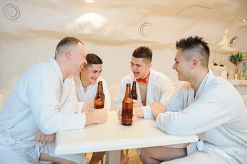Fototapeta na wymiar close up photo of 4 men in white gowns sitting around a table and drinking beer after sauna procedures