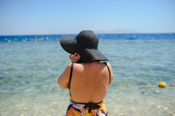 back view of a young woman in swimsuit and a blue big hat sunbathing on the beach