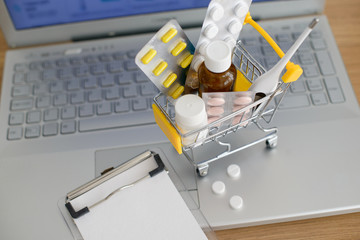 Shopping cart toy with medicaments and paper note in front of laptop screen with pharmacy web site on it. Pills, blister packs, medical bottles, thermometer and recipe set.  - 275741762