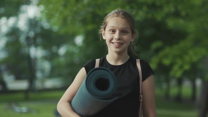 Sweet young Caucasian girl with yoga mat in hands smiling, looking at camera. Portrait of marvelous teenager in sports clothes. Park. Harmonious personality. Outside. Summertime.