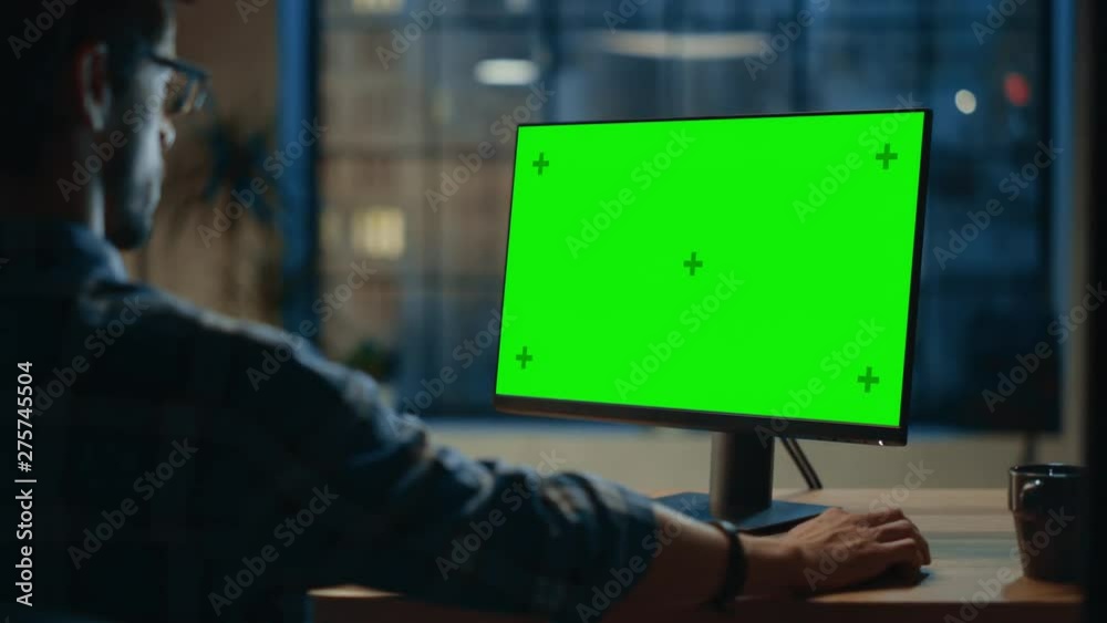 Poster Over the Shoulder: Creative Young Man Sitting at His Desk Using Desktop Computer with Mock-up Green Screen. Evening in the Stylish Office Studio with City Window View