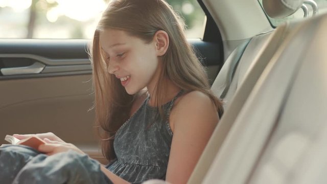 Portrait of smiling cute Caucasian girl holding book in hands. Footage of pretty teenager enjoying reading. Alone in car. Backseat. Summer, daytime. Good habit.