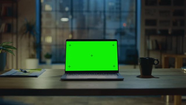 Mock-up Green Screen Laptop Standing on the Desk in the Modern Creative Office. In the Background Warm Evening Lighting and Open Space Studio with City Window View. Zoom in Shot