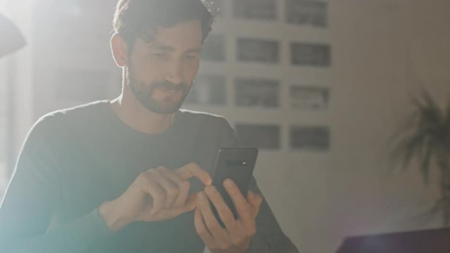 Professional Creative Man Sitting at His Desk in Home Office Studio Using Smartphone Man Using Mobile Phone, Browsing Through Internet, Social Networks / Media, Working on Laptop. Camera Moving Around