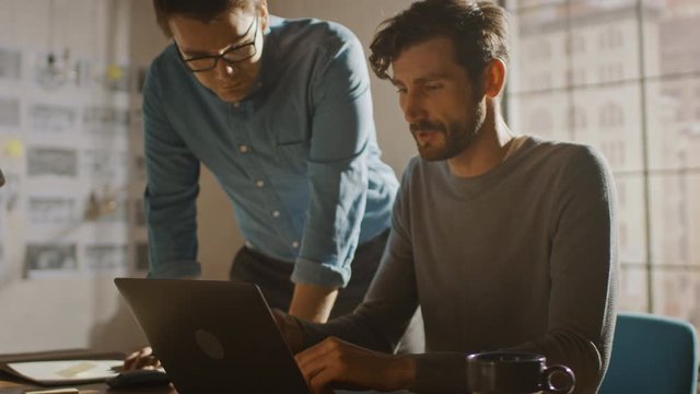 Professional Creative Man Sitting at His Desk works on a Laptop, His Colleague Helps with Poignant Advice, They Have Discussion about Decisions made for Project. 360 Degree Tracking Arc Shot