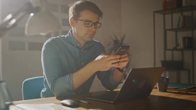 Professional Creative Man Sitting at His Desk in Home Office Studio Working on a Laptop. Man Using Smartphone to Check Social Media, Search Internet. 360 Degree Tracking Arc Shot Movement
