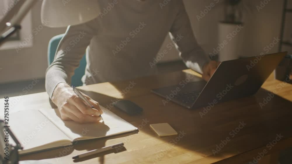 Poster Anonymous Creative Man Sitting at His Desk in Home Office Studio Working on a Laptop Computer, Writes down Notes. Low Angle 360 Degree Tracking Arc Shot Movement. Camera Moving Around