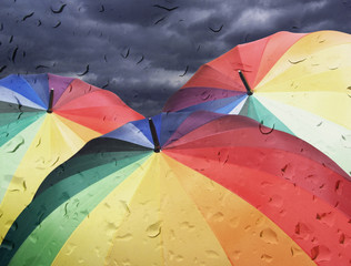 Rainbow umbrellas on dramatic sky background under rain, view through window glass with rain drops