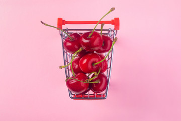 Creative top view of ripe cherries in a shopping cart on pink  background in minimal style. Concept of supermarket or fruit market shopping.