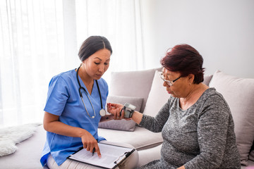 Asian nurse doing blood pressure monitoring for senior woman at home. Close up photo of blood pressure measurement. Nurse measuring blood pressure of senior woman at home. Smiling to each other.