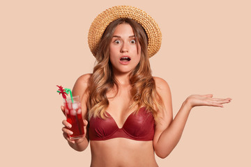 Young european woman wearing red swimming suit and straw hat being shocked due to imminent danger, holding glass of fresh tasty cocktail, posing with open mouth and wide open eyes, expresses astonish.