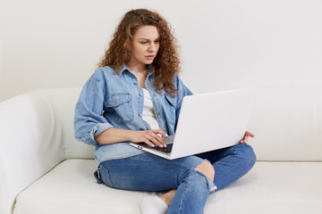 Freelancer female working at home with laptop, has concentrated facial expression, wearing denim jacket and jeans, charming female sitting on white comfortable couch, using wireless internet.