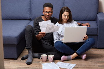 Team work concept. Multiethnic couple working at home, check business plan, looking at documents, sitting on floor near sofa in domestic atmosphere, using laptop computer, wearing casual outfits.