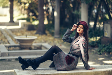 Young beautiful girl wearing winter coat and cap sitting on a bench in urban park.