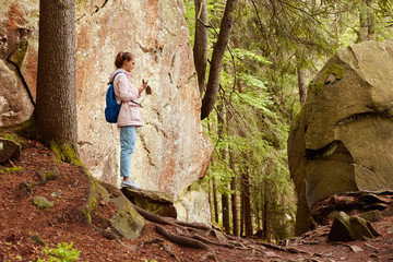 Profile of young attractive tall traveller holding mobile phone in hands, looking attentively at screen, trying to find connection, having orientating problems, taking photos, walking between stones.