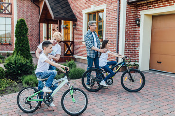 Fototapeta premium Grandparents bike riding with grandchildren. Two smallkids teach to ride a bike near modern house.