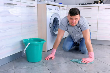 Unhappy tired man in rubber gloves washes the floor in the kitchen and looks at the camera at the end