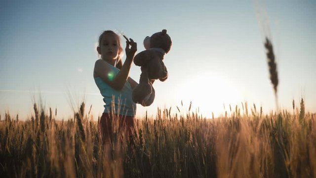 Pretty little girl hugging her teddy bear in wheat field at sunset. Summer time, happy childhood concept.