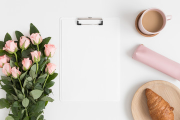 Top view of a white clipboard mockup with a bouquet of pink roses, coffee and a croissant on a white table.
