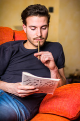 Young man sitting doing a crossword puzzle looking thoughtfully at a magazine, with his pencil to...