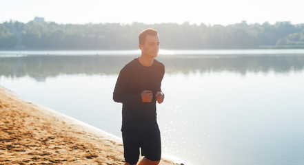 Young man running in the sunlight on the shore of a lake in the morning. Stay fit!