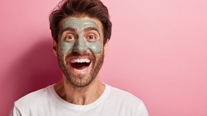Close up portrait of cheerful relaxed man has facial clay mask, receives beauty treatments from professional cosmetologist, wears white t shirt, isolated on pink background, blank space away