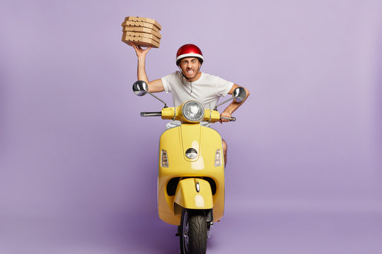 Food service, fast delivery concept. Impatient pizza delivery driver poses on fast motorbike, being in hurry with boxes of fast food, clenches teeth, annoyed with heavy traffic drives to customer home
