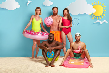 Discontent multiethnic friends spend free time at coast, hold different swimming supplies, beachball, tennis racket, gaze in displeasure, worried by bad situation at beach. Holiday and resort concept