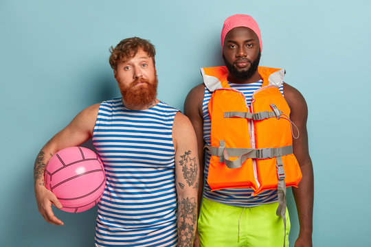 Serious multiethnic men wear casual clothes, swimhat, lifejacket, play acive games at beach with ball, feel relaxed and satisfied, stand against blue background. Diverse tourists spend summer holiday
