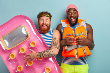 Excited overemotive two men gaze happily, spend free time near water, hold pink mattress for floating, prepare for pool party, have fun during summer day. Multiethnic vacationists enjoy swimming