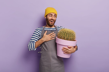 Wow, what great cactus I grew! Cheerful unshaven male botanist looks gladfully at potted plant, enjoys gardening, rejoices awesome results of fertilizing, keeps palm on chest from excitement © WHstudio Leushin N