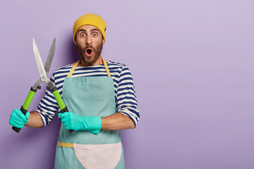 Astonished male gardener carries pruning shears, going to cut green big bush, works in own garden, surprised with much work, wears rubber protective gloves and apron, stands indoor over purple wall