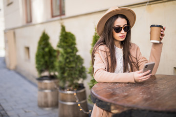Young pretty woman in a coat and hat sits in a street cafe at a table with a glass of coffee use phone and waiting for a date of friends or a guy