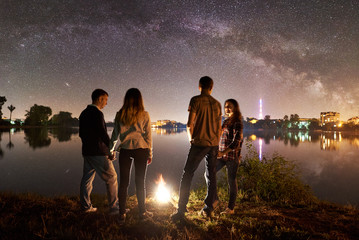 Two couple having a rest on lake shore near bonfire together. Friends enjoying beautiful view of night sky full of stars and Milky way, quiet water surface and luminous town on background.