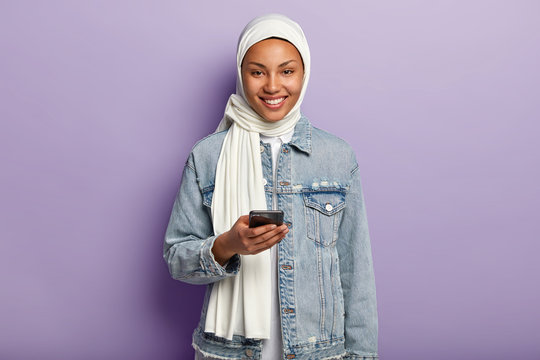 Portrait Of Happy Muslim Lady Wears White Hijab On Head According To Traditions, Uses Modern Cell Phone, Sends Messages, Checks Information Online, Isolated On Purple Wall. Modern Technologies