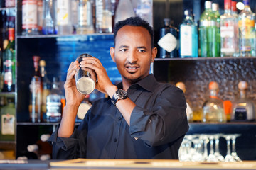 Professional african bartender male holding in hands a shaker with a fresh delicious cocktail. Bartender shaking a cocktail shaker as she stands behind the bar mixing a drink for a client. Close-up
